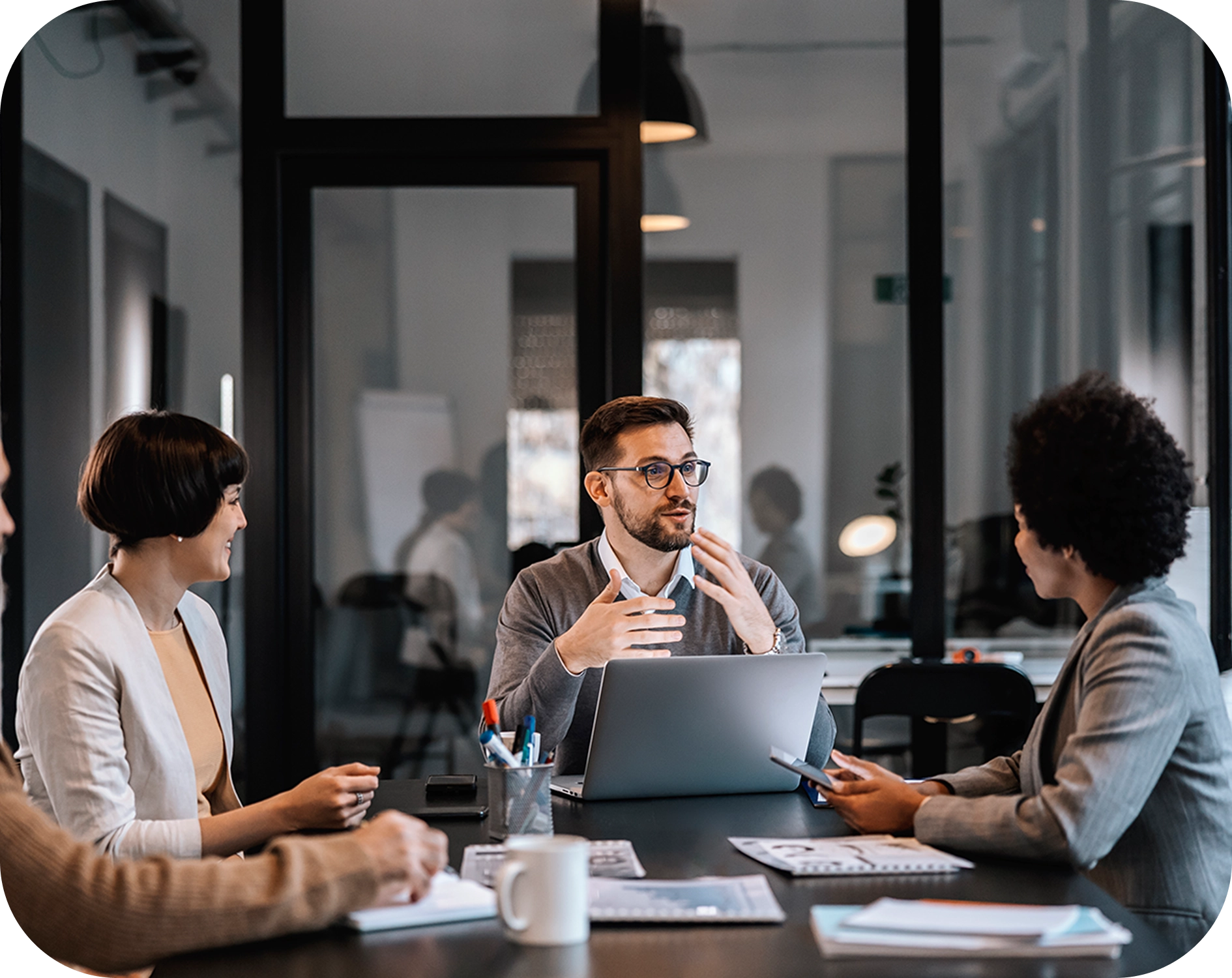 Four people in a modern office have a discussion around a table with a laptop, documents, and coffee mugs.