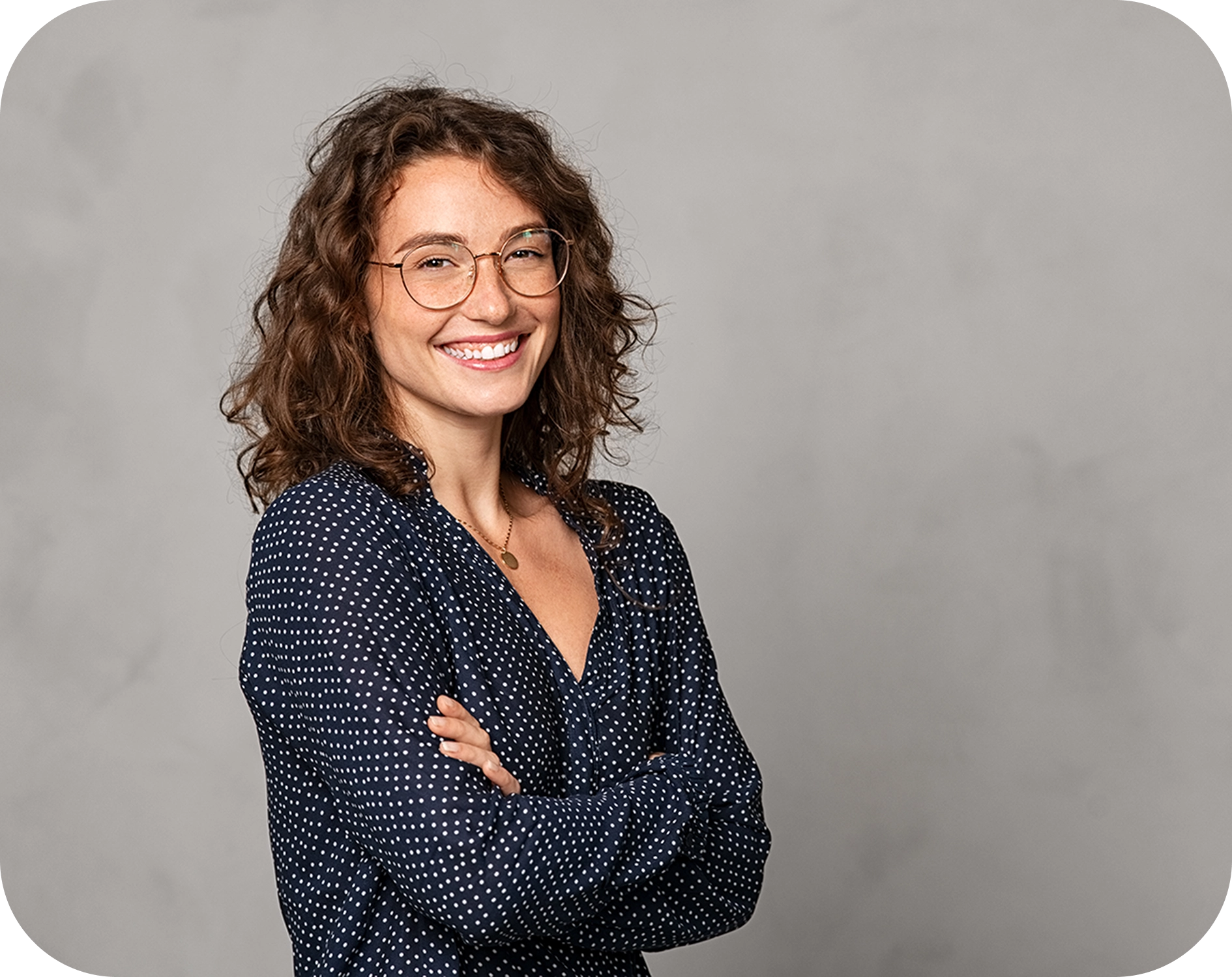 A woman with curly hair and glasses smiles with her arms crossed, wearing a navy polka dot blouse, against a plain gray background.