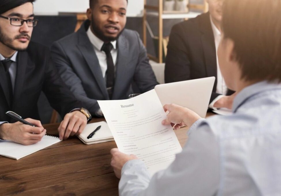 Person sitting at a table facing three individuals in suits, holding a resume.