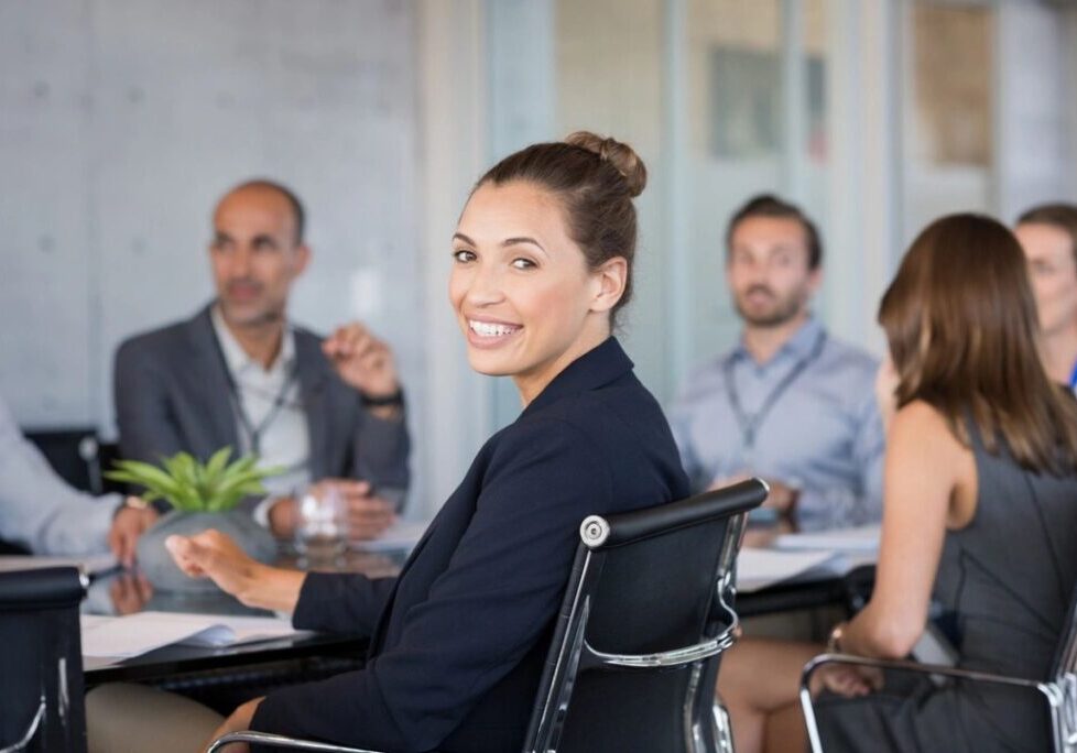 A woman in a business meeting turns to smile at the camera, with several colleagues seated around a conference table.